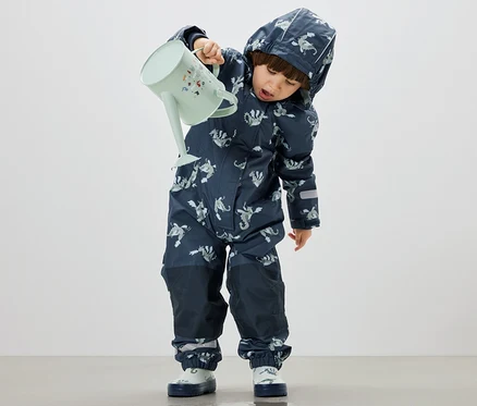 Junge gießt Wasser aus einer pastellgrünen Gießkanne auf den Boden, trägt dabei einen Kinder-Regen-Einteiler, Ritter.