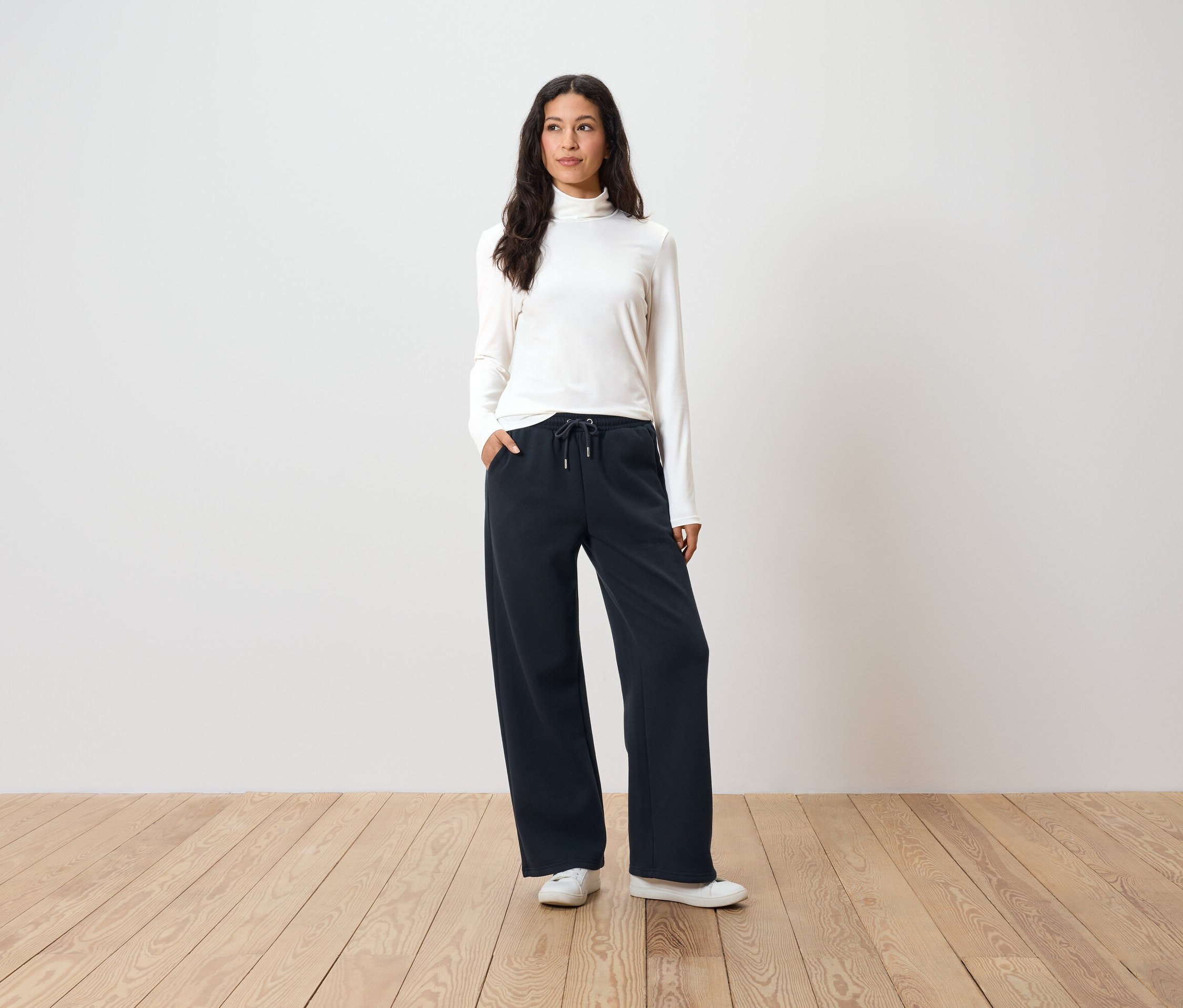 Frau steht mit Händen in den Taschen einer dunkelblauen, weiten Hose, einem weißen Rollkragenpullover und weißen Turnschuhen.
