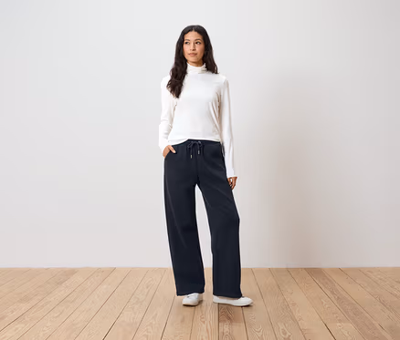 Frau steht mit Händen in den Taschen einer dunkelblauen, weiten Hose, einem weißen Rollkragenpullover und weißen Turnschuhen.