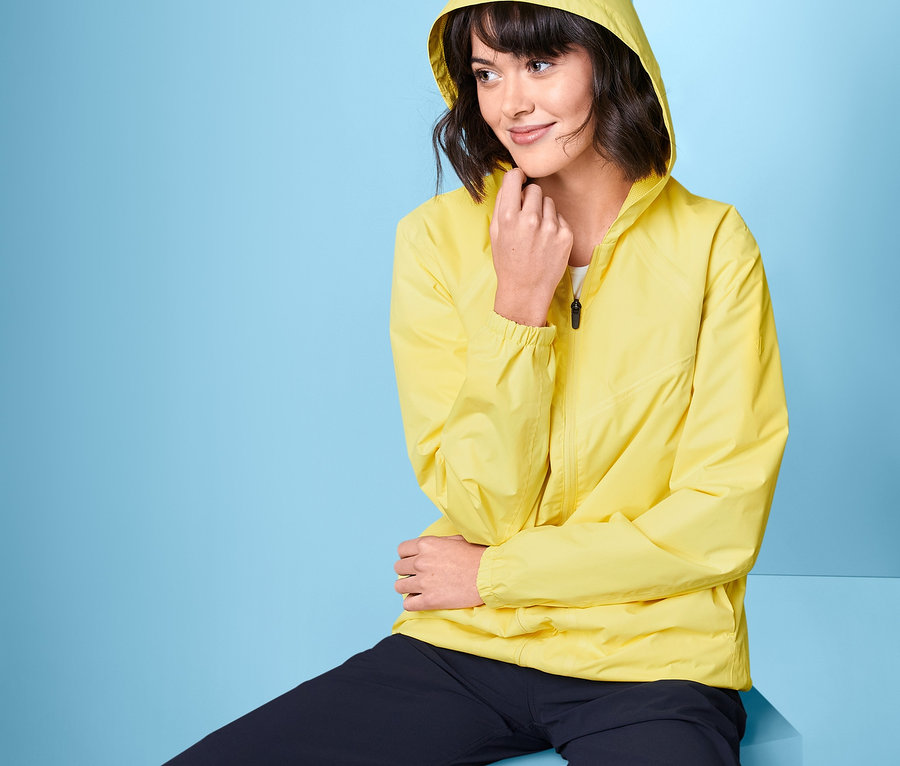 Brünette sitzt auf einem blauen Würfel und trägt eine gelbe Lightweight-Regenjacke »pack me«.