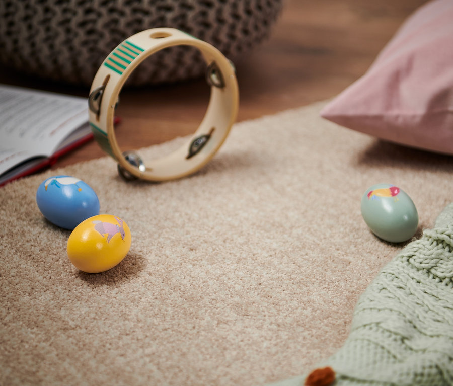 Auf einem beigen Teppich liegen ein offenes Buch, ein Tamburin und drei Rassel-Eier.