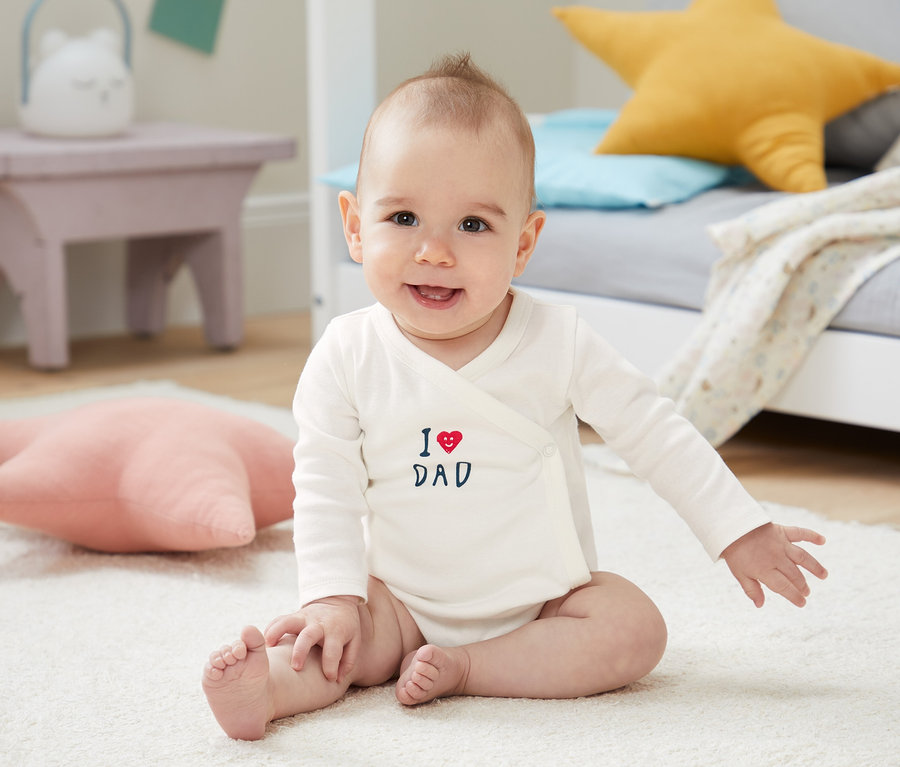 Lächelndes Baby sitzt auf einem weißen Teppich und trägt einen weißen 5er Langarm-Body aus Bio-Baumwolle mit der Aufschrift "I love Dad".