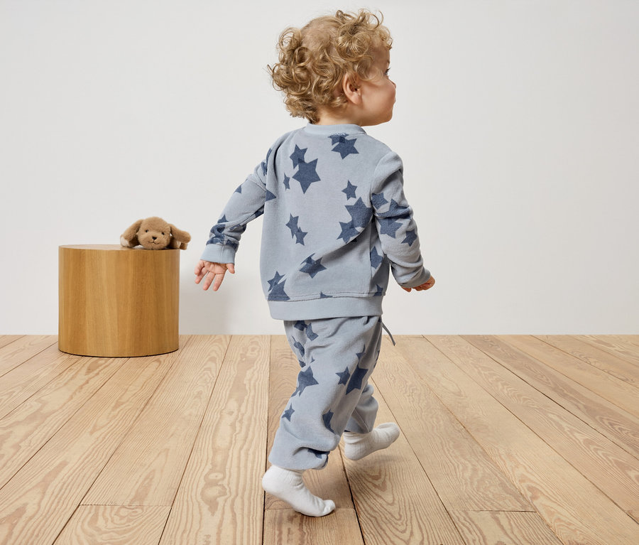 Kleinkind in blauer Sternenkleidung geht auf Holzboden. Auf dem Hocker liegt ein Plüschhund.