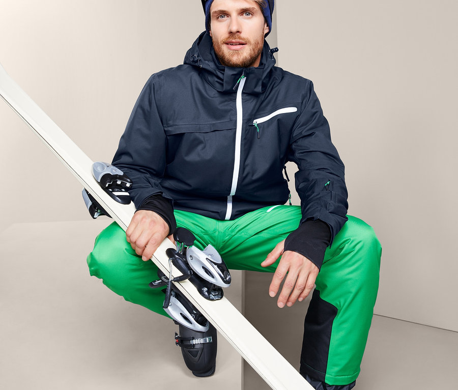 Mann mit Skijacke, Skibrille und grüner Hose hält Ski.