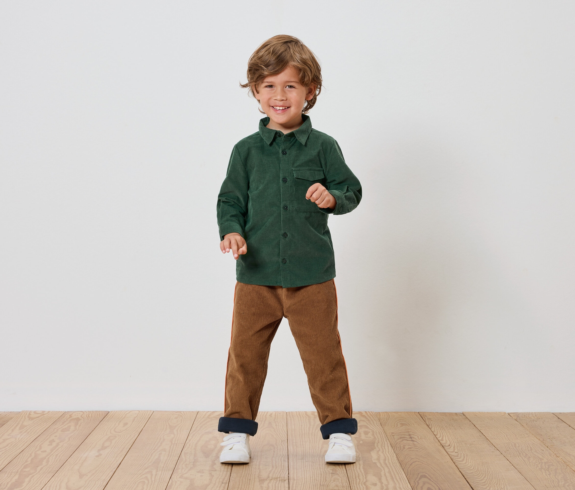 Junge steht und lächelt, trägt ein grünes Hemd, braune Hosen und weiße Schuhe.