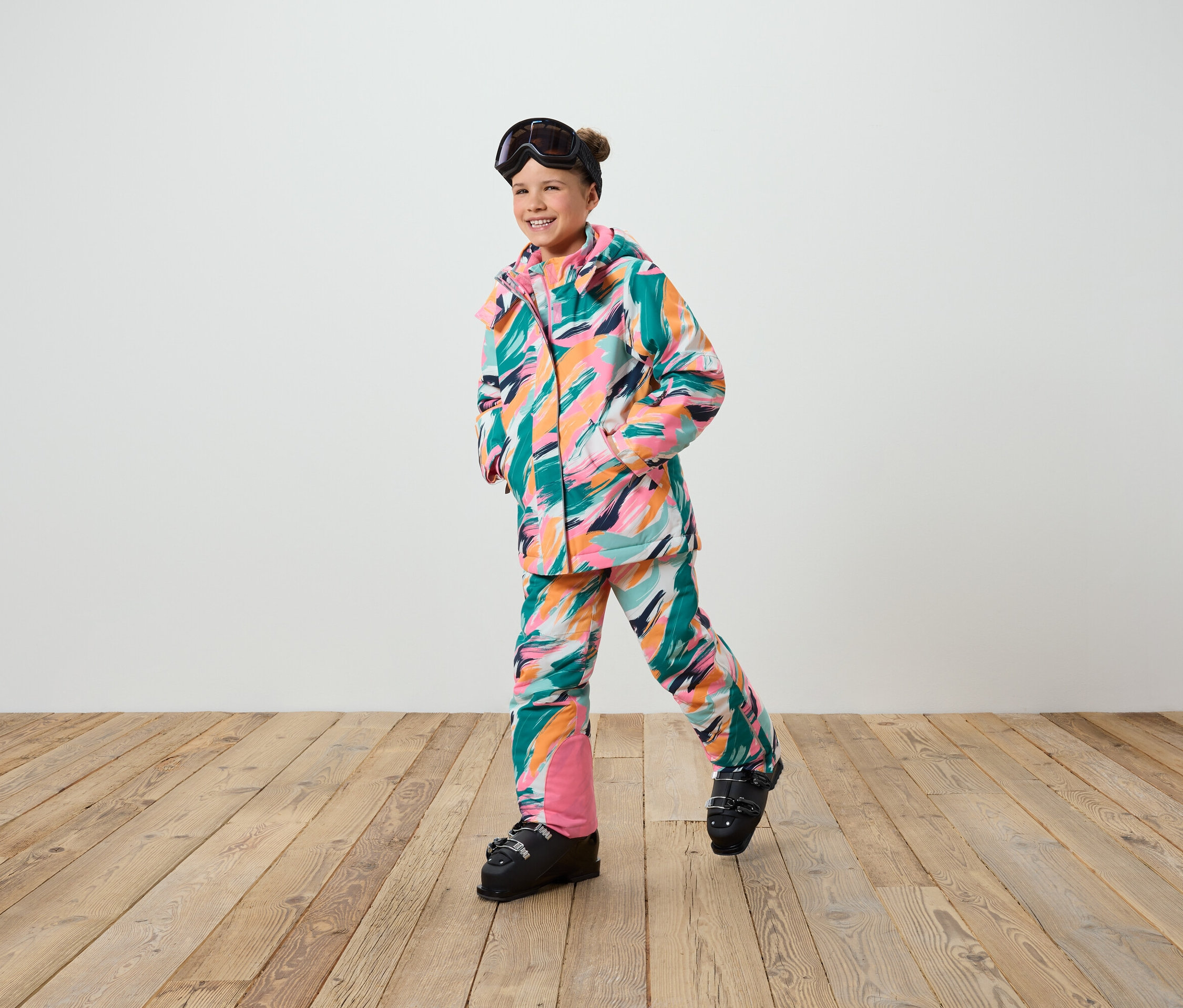 Lächelndes Mädchen posiert in einem Skianzug mit bunten Strichen und Skischuhen.