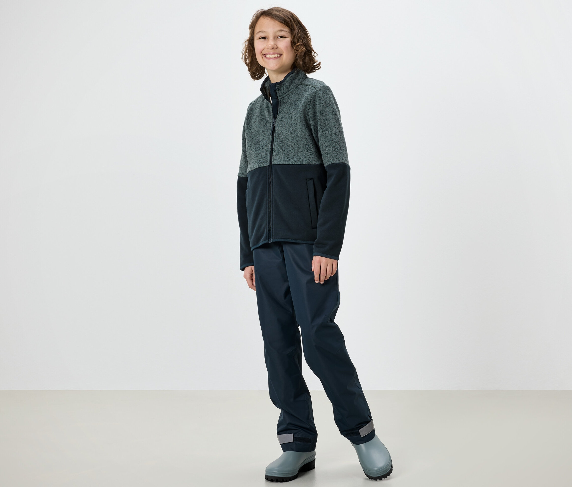 Kind posiert in Hybrid-Strickfleecejacke, Hose und Gummistiefeln.