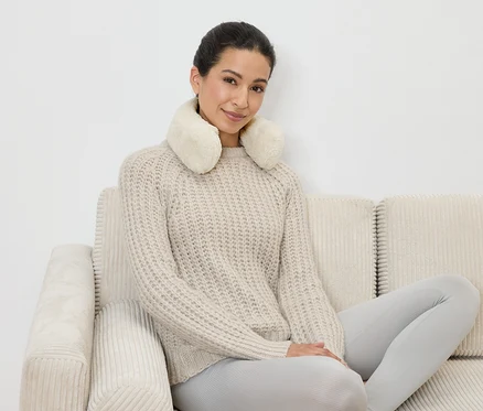 Frau sitzt auf einem beigen Sofa mit einem cremefarbenen Entspannungskissen um den Nacken, trägt einen Pullover und Leggings.