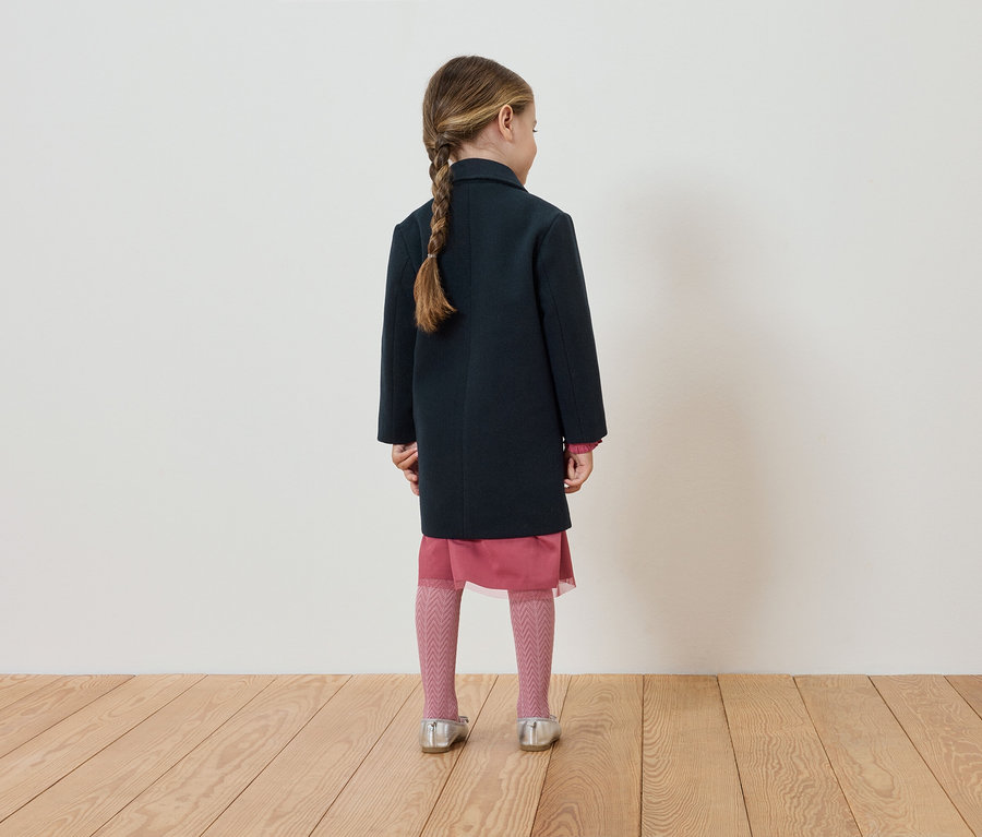 Ein Mädchen mit Zopf von hinten trägt einen dunkelblauen Mantel, ein rosa Kleid, Strumpfhosen und silberne Schuhe.