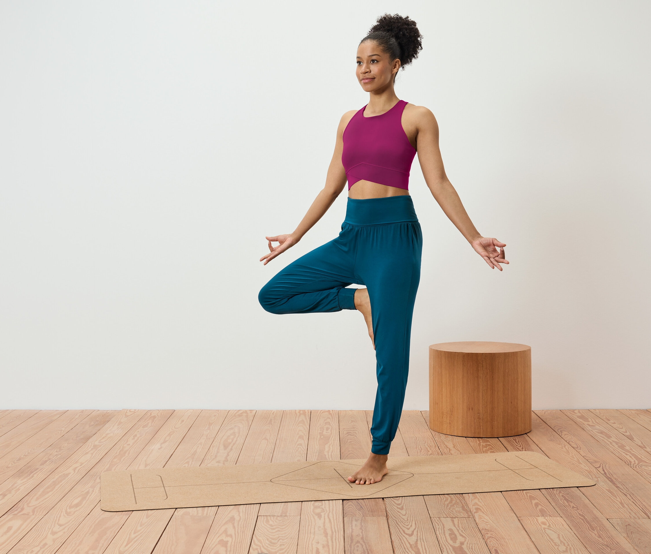 Frau in pinkem Top und blauer Hose steht in der Baumposition auf einer Yogamatte.