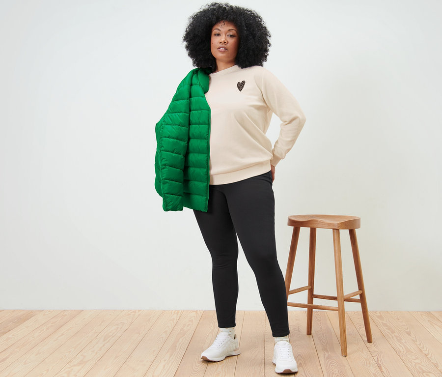 Frau posiert in einem cremefarbenen Sweatshirt mit Print, schwarzen Leggings und weißen Schuhen. Eine grüne Jacke hängt über ihrer Schulter.