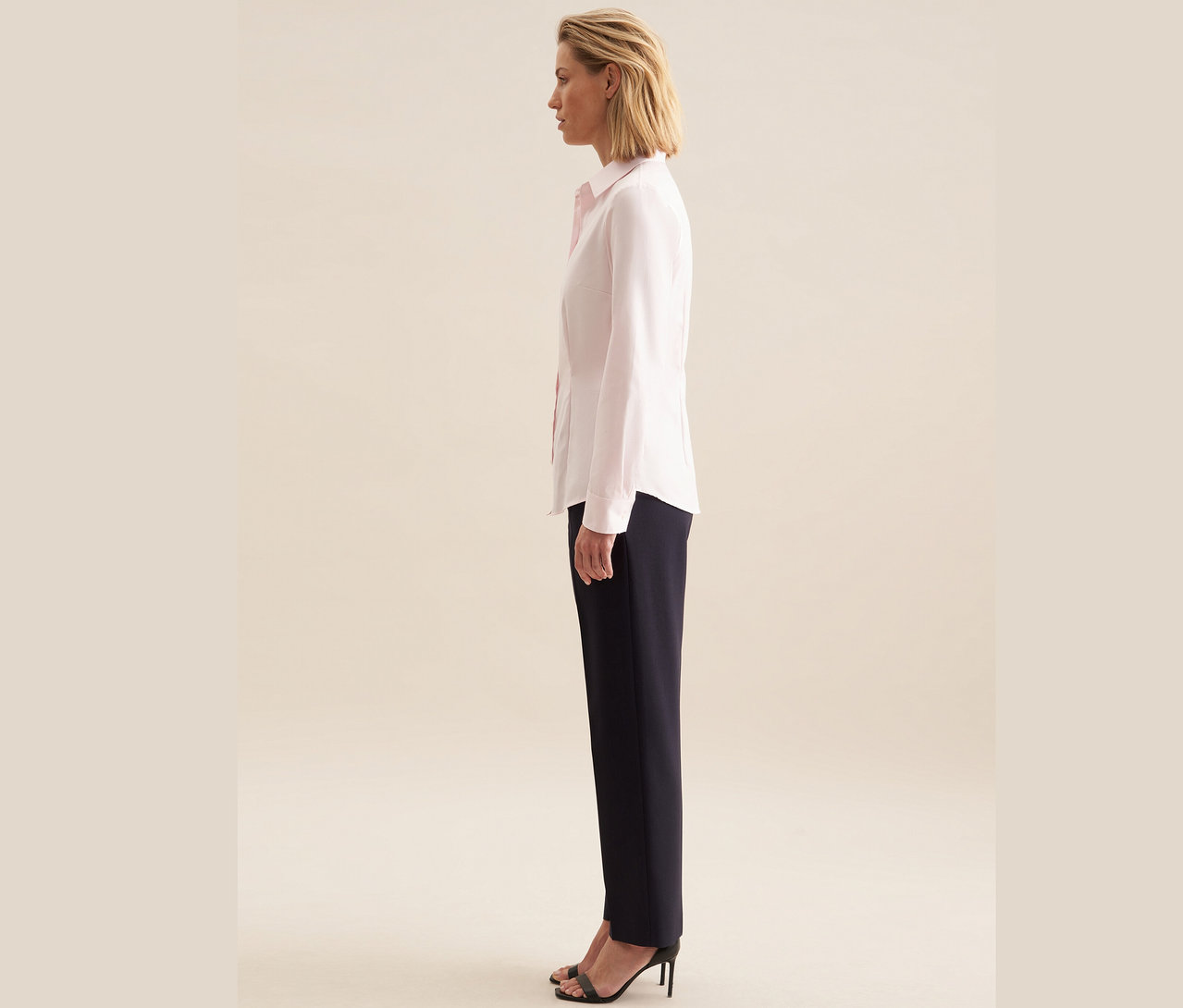 Frau steht seitlich und trägt eine roséfarbene Seidensticker Twillbluse, dunkelblaue Hose und schwarze Riemchensandalen mit Absatz.