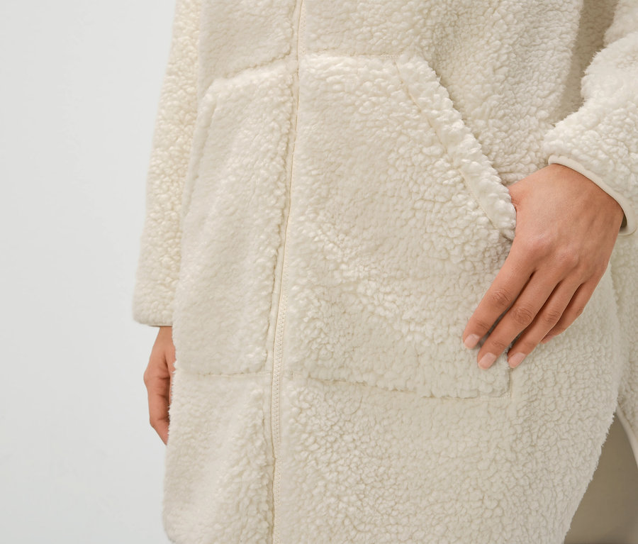 Detailaufnahme eines cremefarbenen Damen-Bademantels aus Teddyfell mit Reißverschluss und Taschen, Hand in der Tasche.