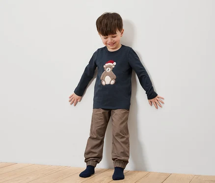 Junge steht vor einer weißen Wand und trägt ein dunkelblaues Hemd mit einem Bären mit Weihnachtsmütze und braune Hosen.