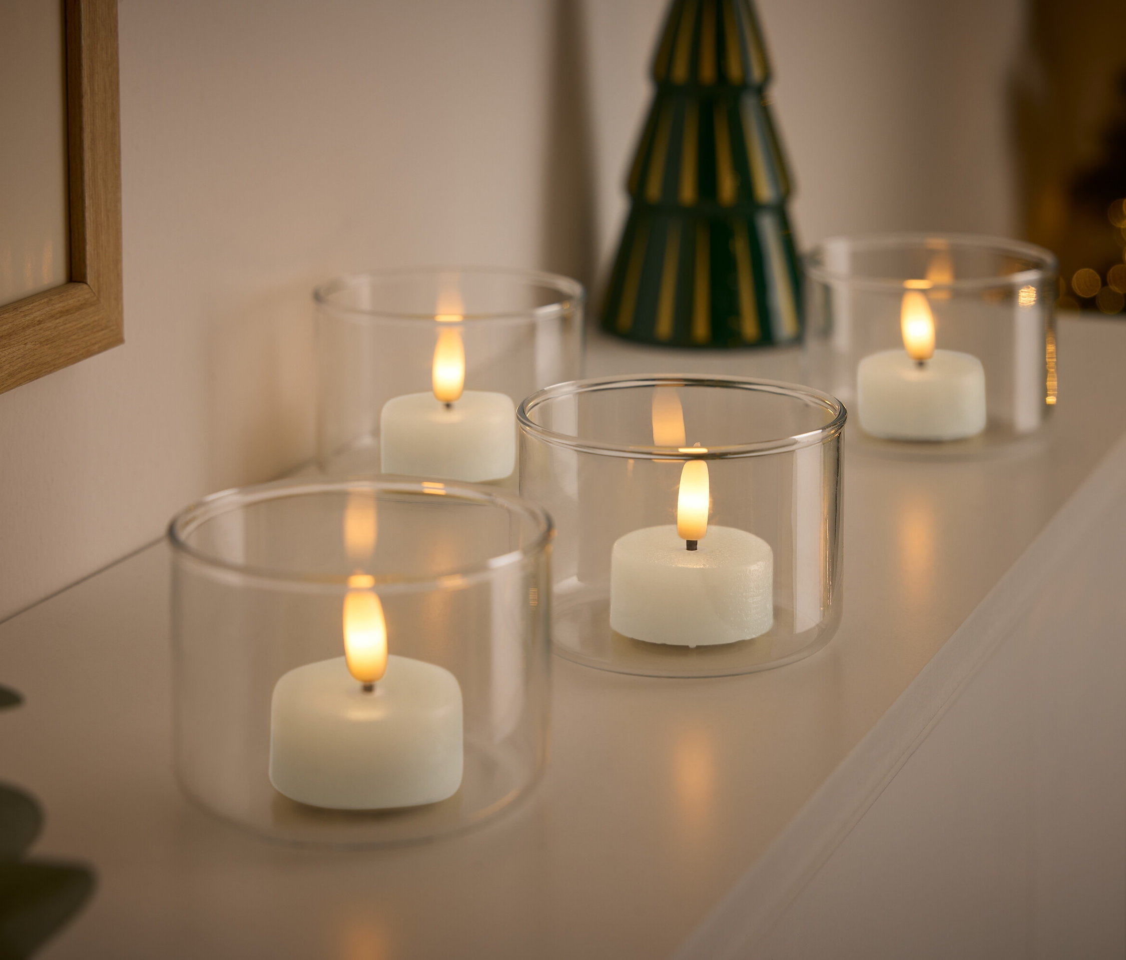 Vier weiße Kerzen in durchsichtigen Glasbehältern mit einem kleinen grünen Weihnachtsbaum im Hintergrund.