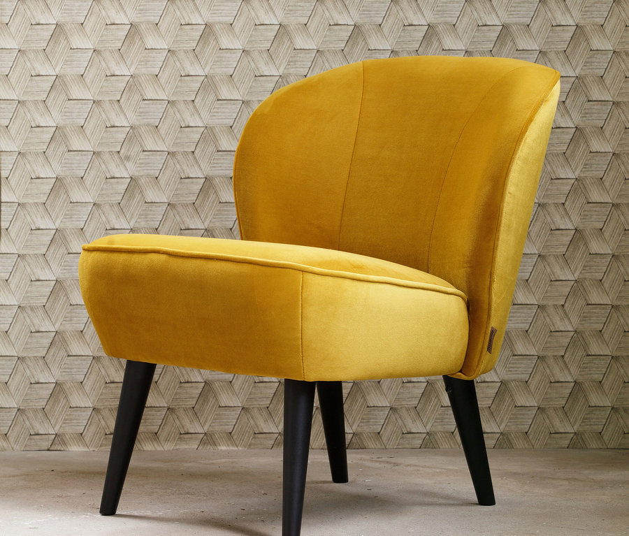 Ein ockerfarbener Woood Sessel "Sara" steht vor einer Wand mit geometrischem Muster.