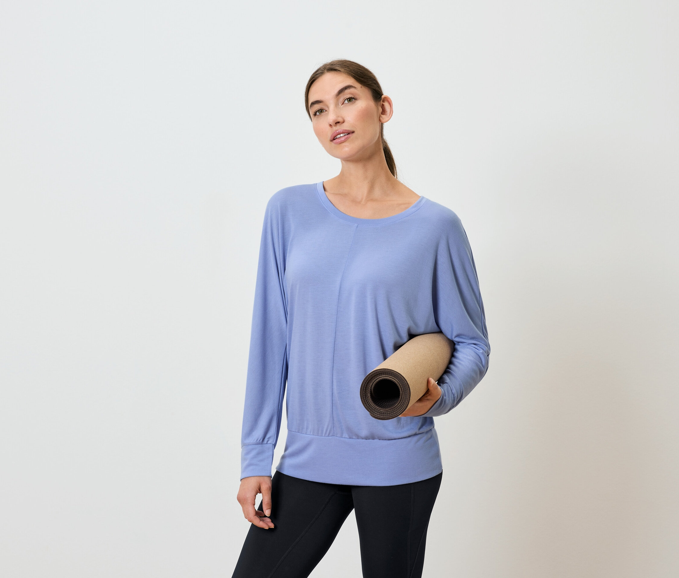 Frau hält eine zusammengerollte Yogamatte, trägt ein blaues Langarmshirt und schwarze Leggings.
