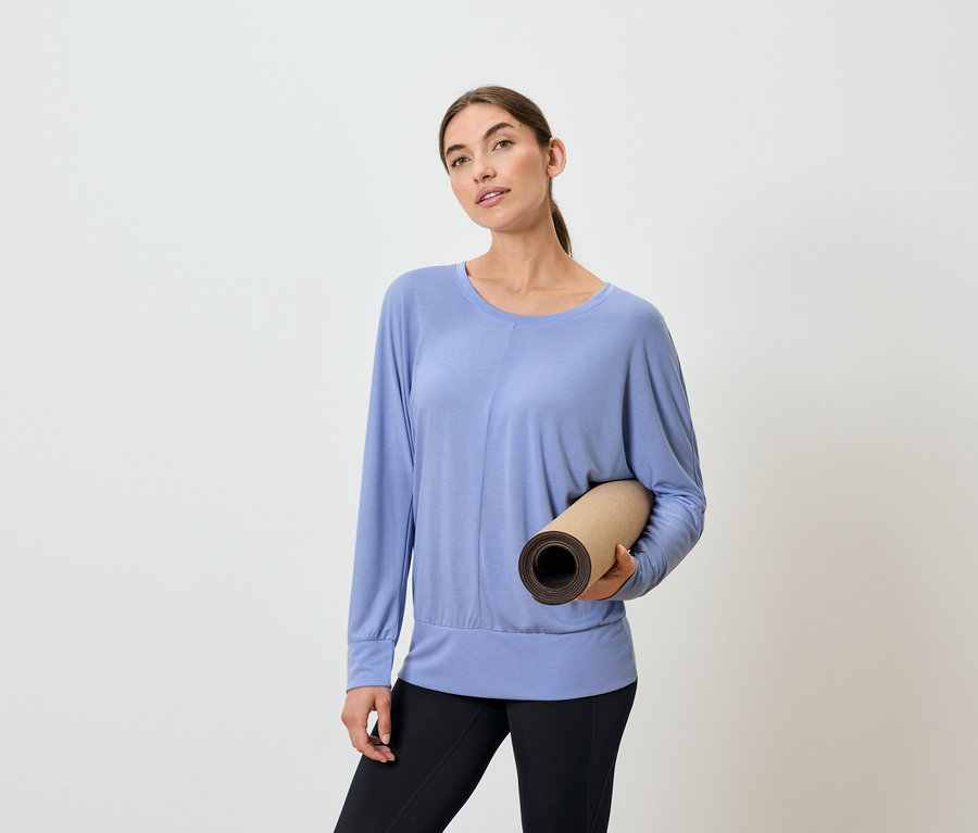 Frau hält eine zusammengerollte Yogamatte, trägt ein blaues Langarmshirt und schwarze Leggings.
