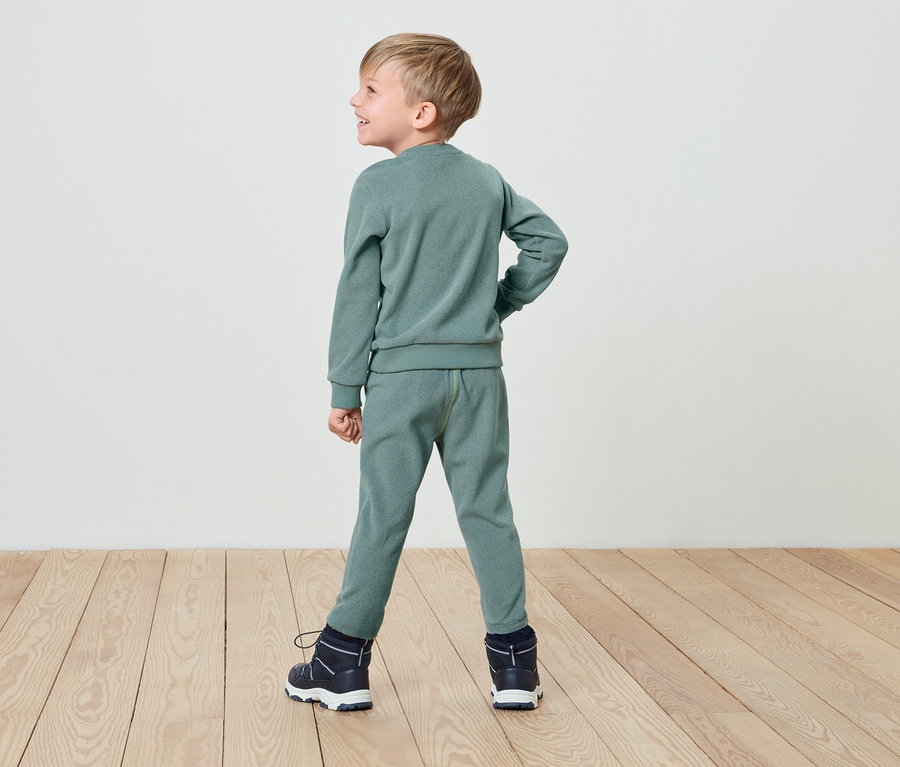 Junge in grünem Trainingsanzug und Stiefeln steht mit dem Rücken zur Kamera auf einem Holzboden.