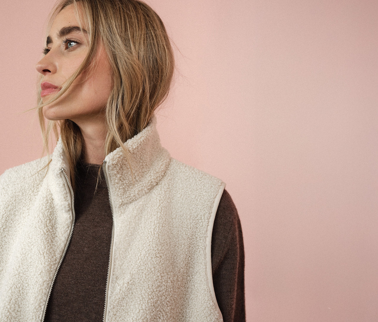 Frau trägt eine Street One Teddyweste über einem braunen Pullover, Blickrichtung nach links vor einem rosa Hintergrund.