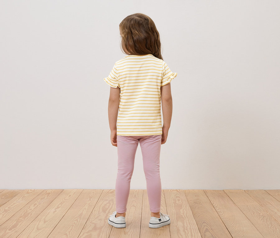 Mädchen steht mit dem Rücken zur Kamera, trägt ein gelb-weiß gestreiftes Oberteil mit Rüschen, rosa Leggings und weiße Turnschuhe.