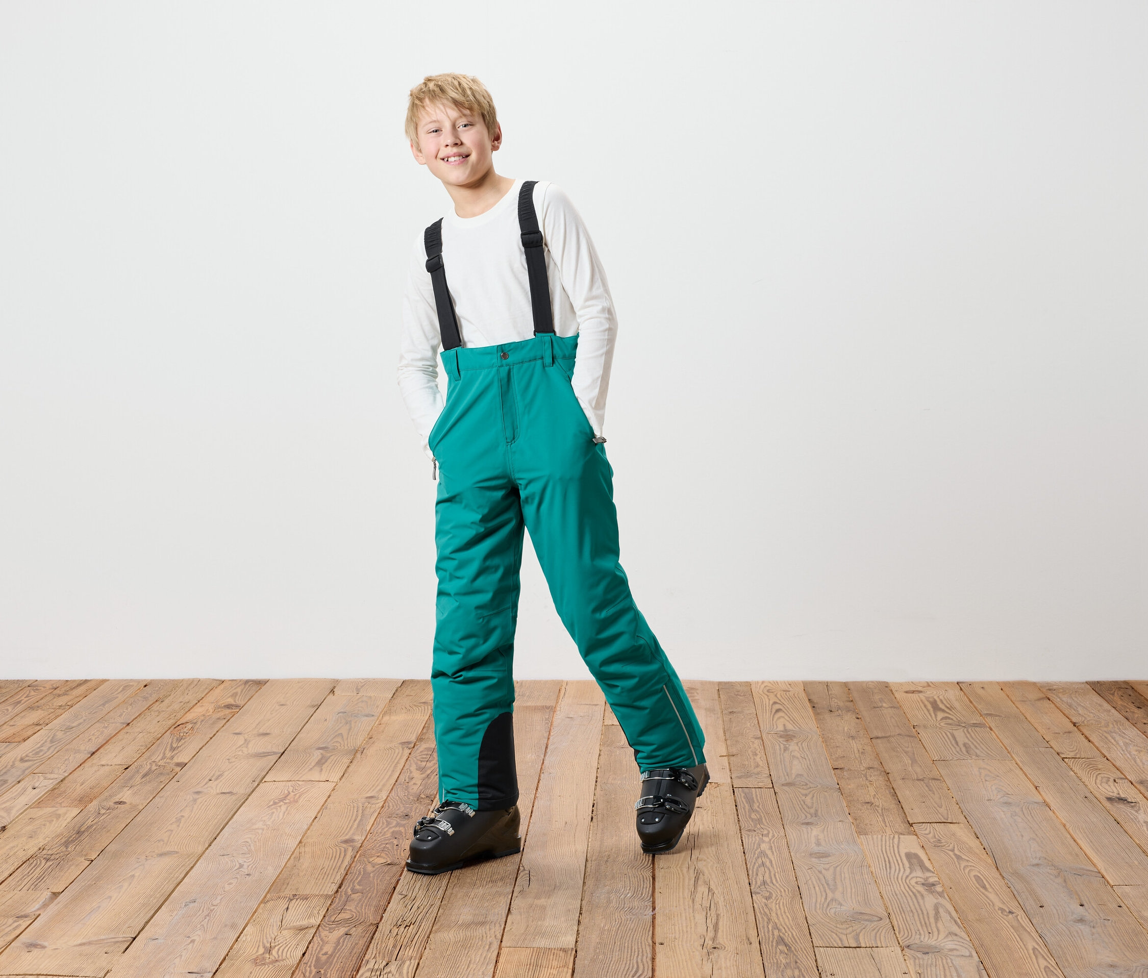 Ein Junge in grüner Skihose mit schwarzen Hosenträgern und Skischuhen steht auf einem Holzboden vor einer weißen Wand.