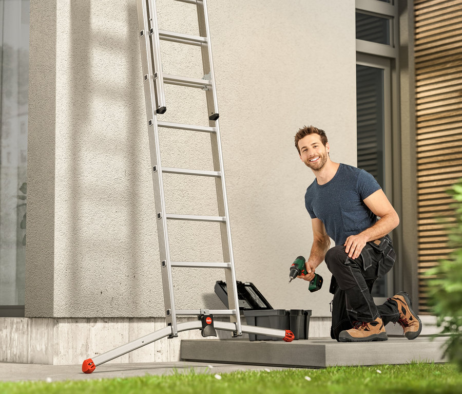 Mann kniet neben einer Leiter und hält einen Bohrer in der Hand.