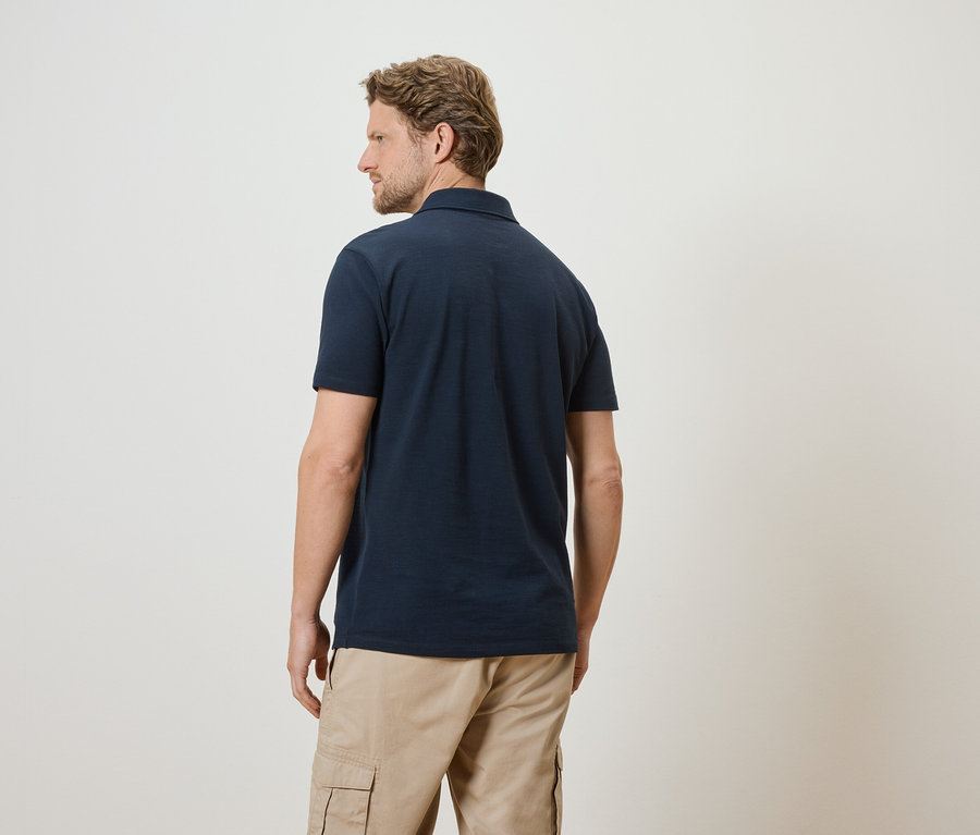 Mann von hinten in marineblauem Poloshirt und beigen Cargohosen vor weißem Hintergrund.