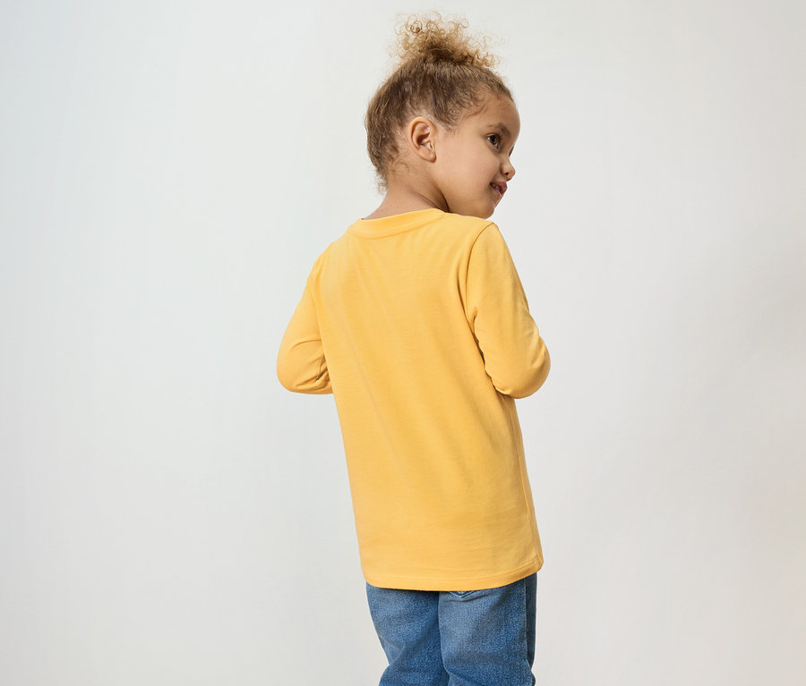 Ein junges Mädchen mit lockigem Haar trägt ein gelbes Langarmshirt und Jeans.