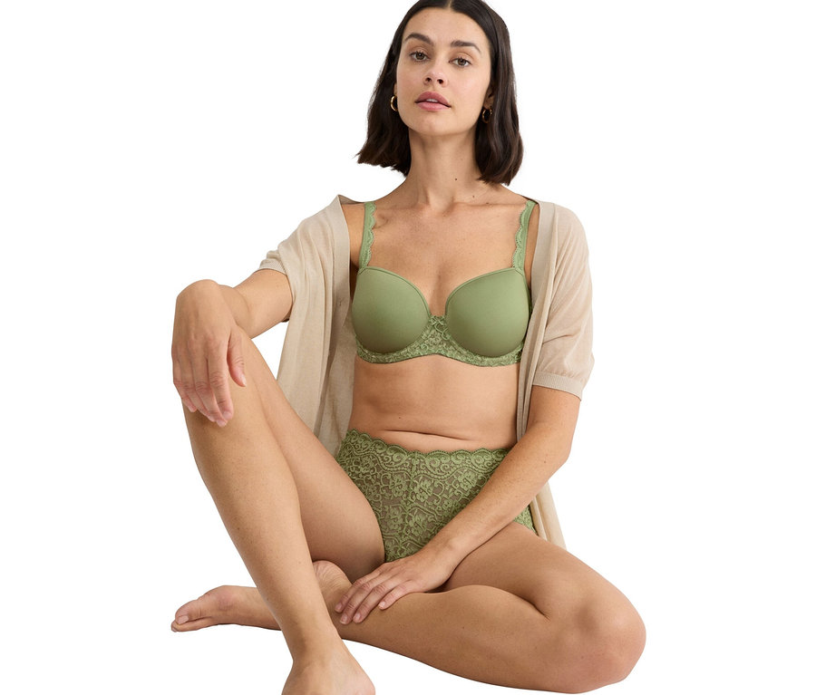 Frau sitzt mit gekreuzten Beinen und trägt einen grünen BH und eine grüne Spitzhose, zusammen mit einer beigen Strickjacke.