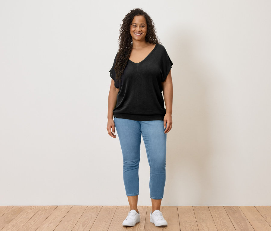 Eine Frau mit lockigem Haar steht vor einer Wand und trägt ein schwarzes Oberteil, Jeans und weiße Schuhe.