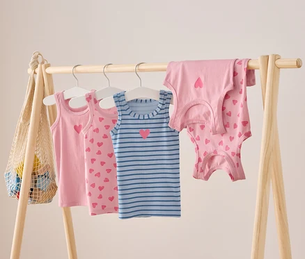 An einer Holzstange hängen rosa und blau gestreifte Kinder Tank Tops mit Herz Motiven.