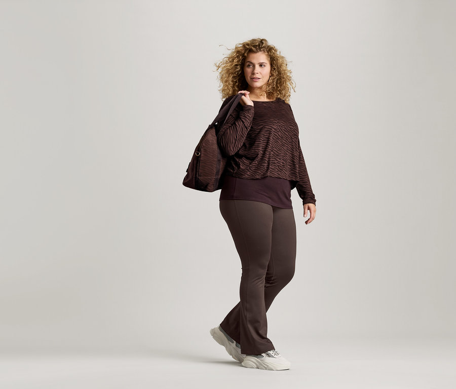 Frau mit lockigen Haaren trägt ein braunes Zebra-Print-Oberteil, braune Leggings und Turnschuhe. Sie hat eine braune Tasche über der Schulter.