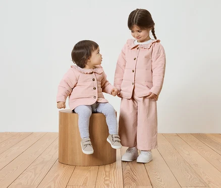 Zwei Mädchen, eines sitzt auf einem Holzhocker, beide tragen rosa Jacken mit Rüschenkragen.