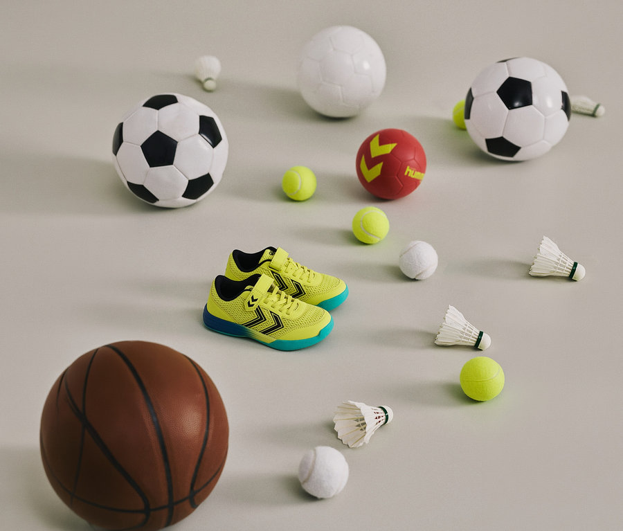 Gelbe hummel® Multiplay Flex VC JR Schuhe zwischen Bällen und Federbällen auf hellem Grund.