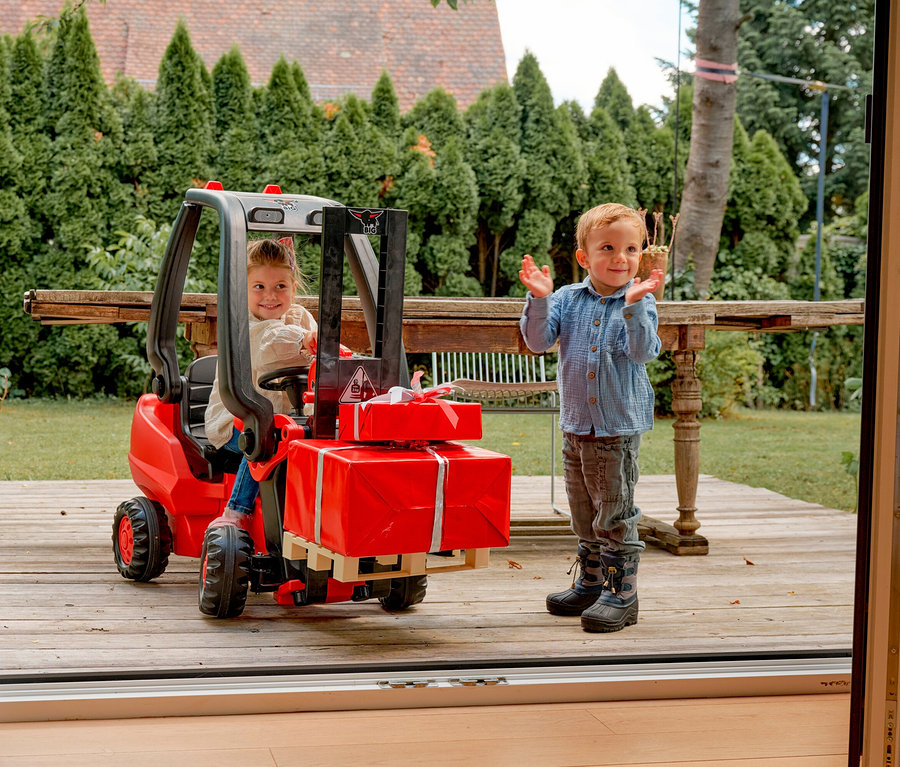 Mädchen fährt mit BIG Tretfahrzeug »Linde Gabelstapler«, während Junge daneben steht und klatscht. Beide sind im Garten auf einer Holzterrasse.