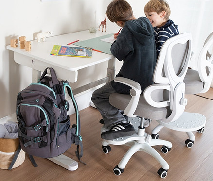 Zwei Kinder sitzen an einem weißen Schreibtisch und zeichnen. Ein grauer Rucksack hängt am Schreibtisch. Beide sitzen auf weißen Bürostühlen.