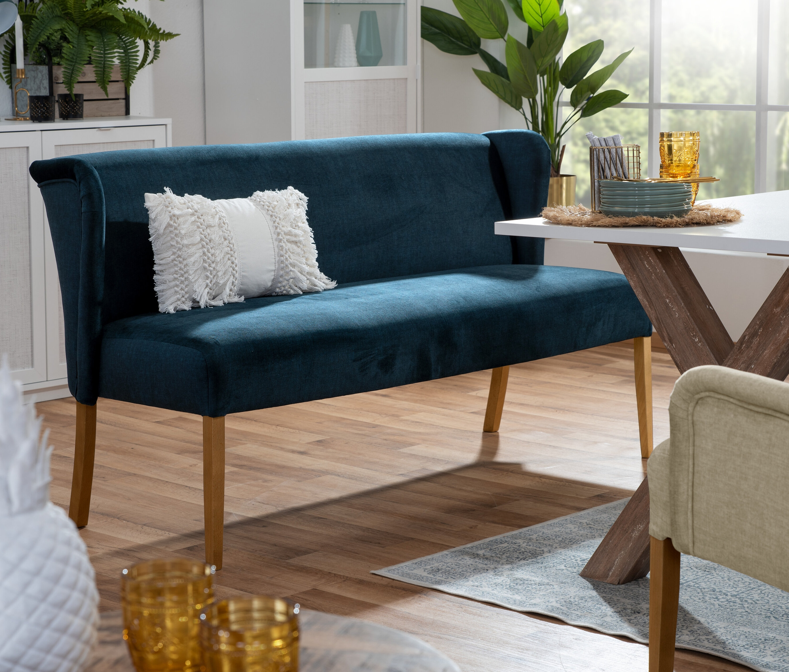 Im Esszimmer steht eine dunkelblaue Polsterbank »Hendrick« mit weißem Kissen vor einem Esstisch mit Geschirr.