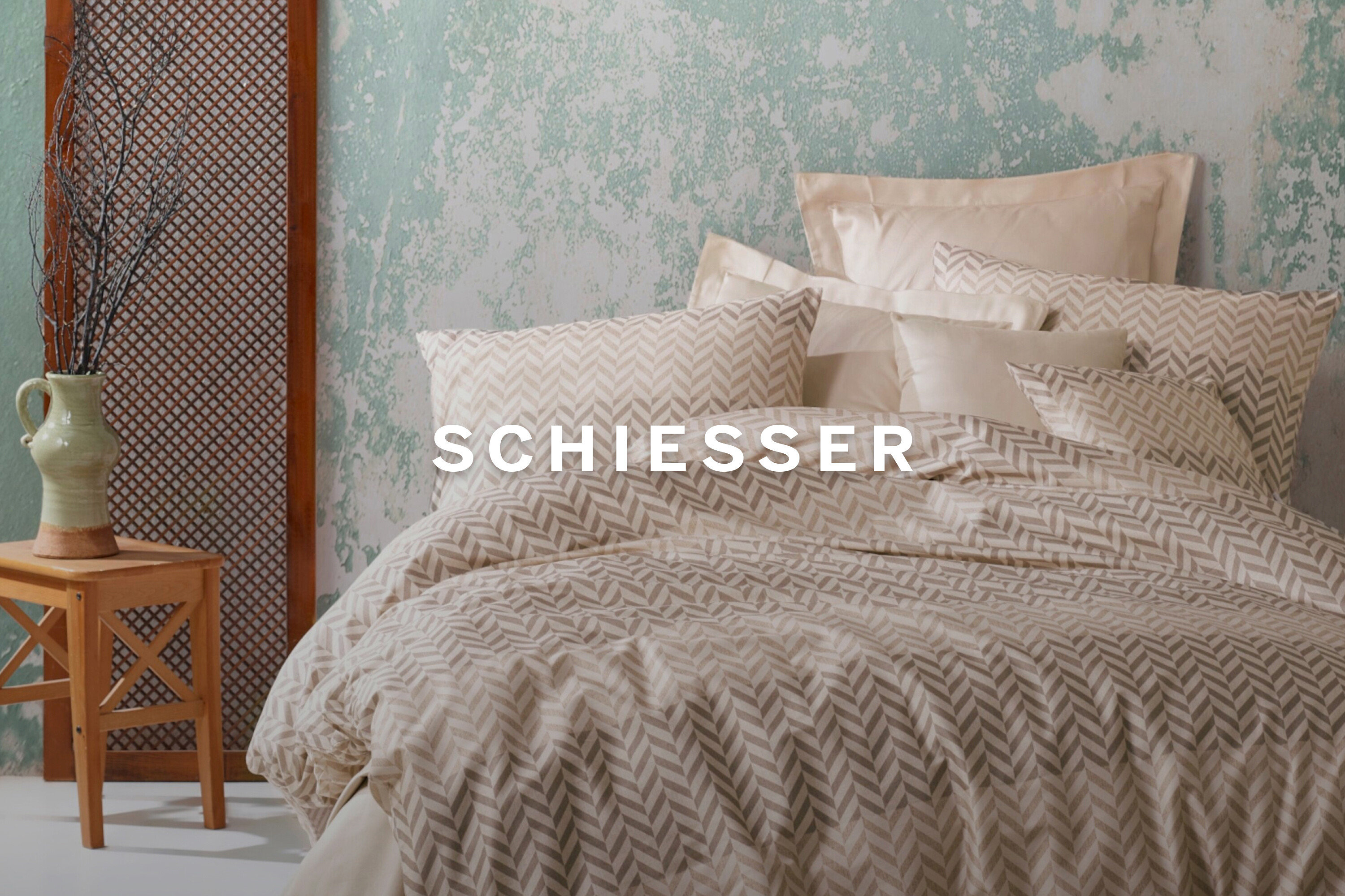 Bett mit Kissen und Decke mit Fischgrätenmuster. Vase mit Zweigen auf einem Holzhocker neben einem Raumteiler.