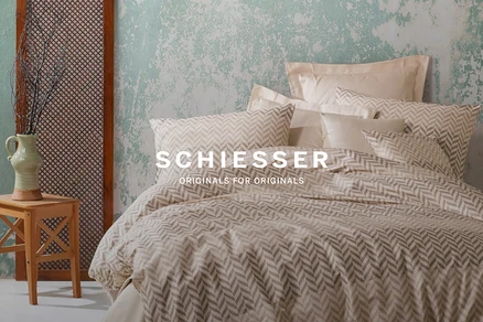 Bett mit Kissen und Decke mit Fischgrätenmuster. Vase mit Zweigen auf einem Holzhocker neben einem Raumteiler.