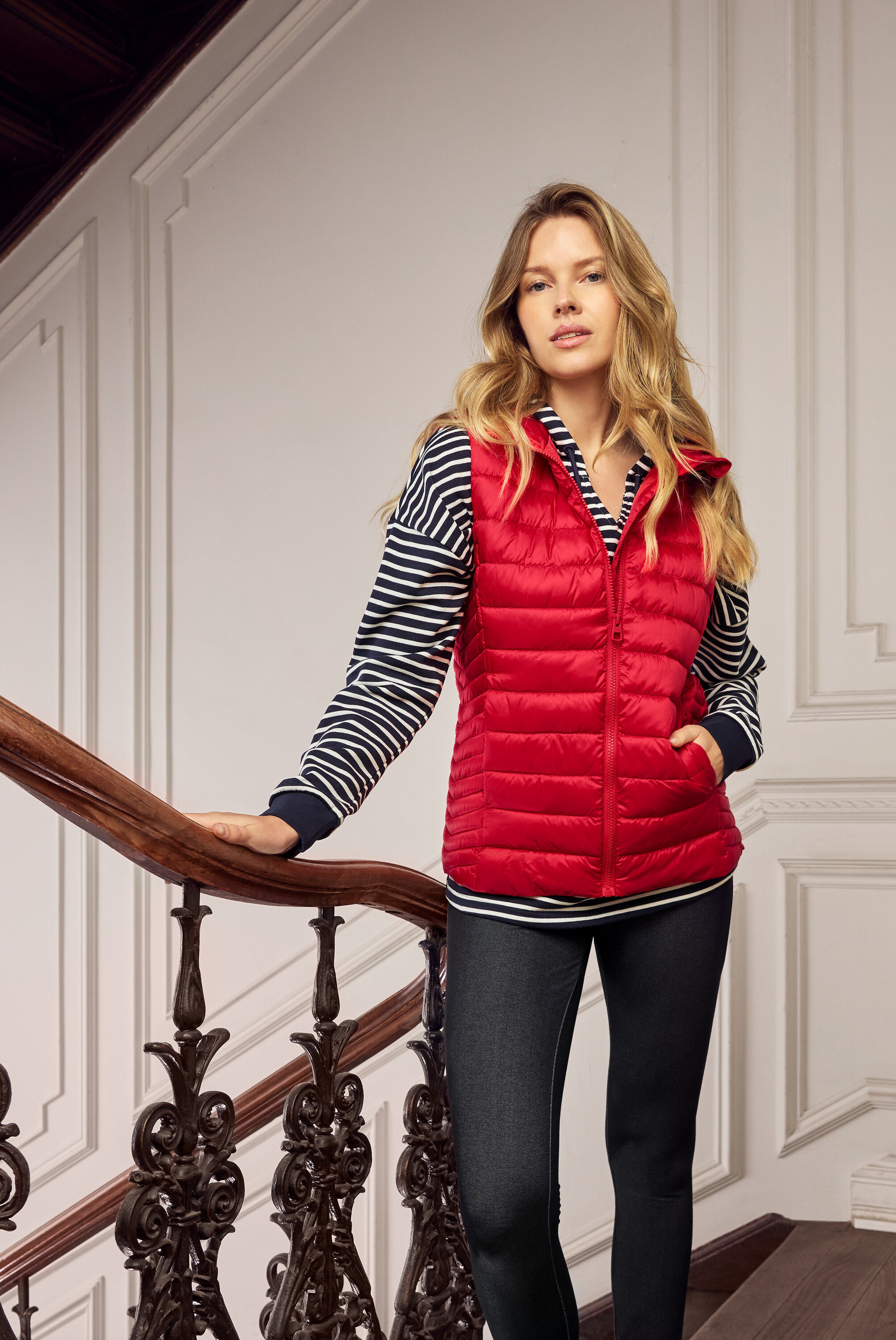 Frau mit blonden Haaren, die eine rote Steppweste, ein blau-weiß gestreiftes Oberteil und dunkle Jeans auf einer Treppe trägt.