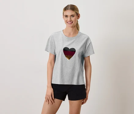 Frau trägt ein graues T-Shirt mit einem Herz in den Farben Schwarz, Burgunderrot und Gold und schwarze Shorts.
