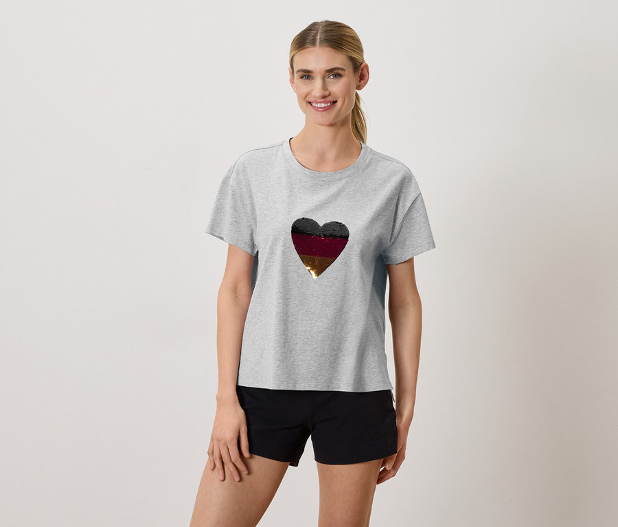 Frau trägt ein graues T-Shirt mit einem Herz in den Farben Schwarz, Burgunderrot und Gold und schwarze Shorts.