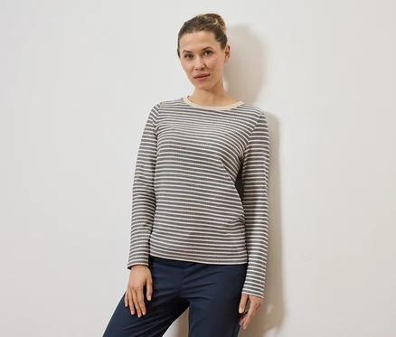 Frau mit Dutt trägt ein grau-weiß gestreiftes Langarmshirt und eine dunkelblaue Hose vor einer weißen Wand.