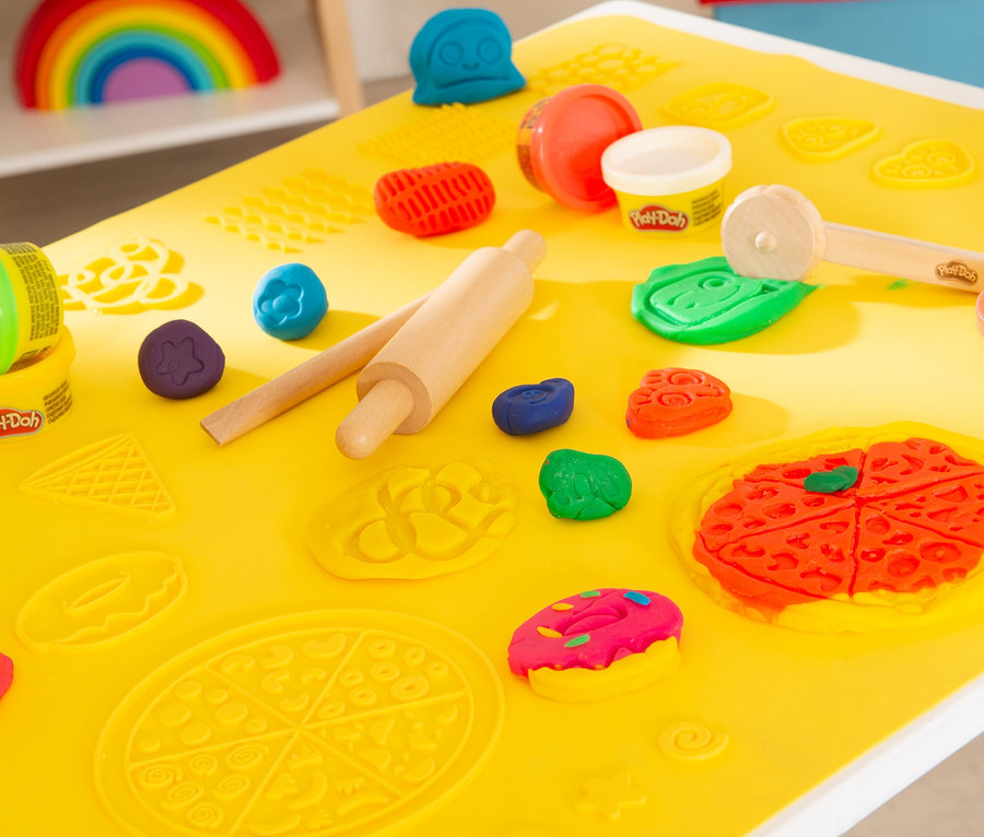 Auf einer gelben Matte liegen Play-Doh, eine Walze und Förmchen. Aus Play-Doh sind Pizza, Donut und Gesicht geformt.