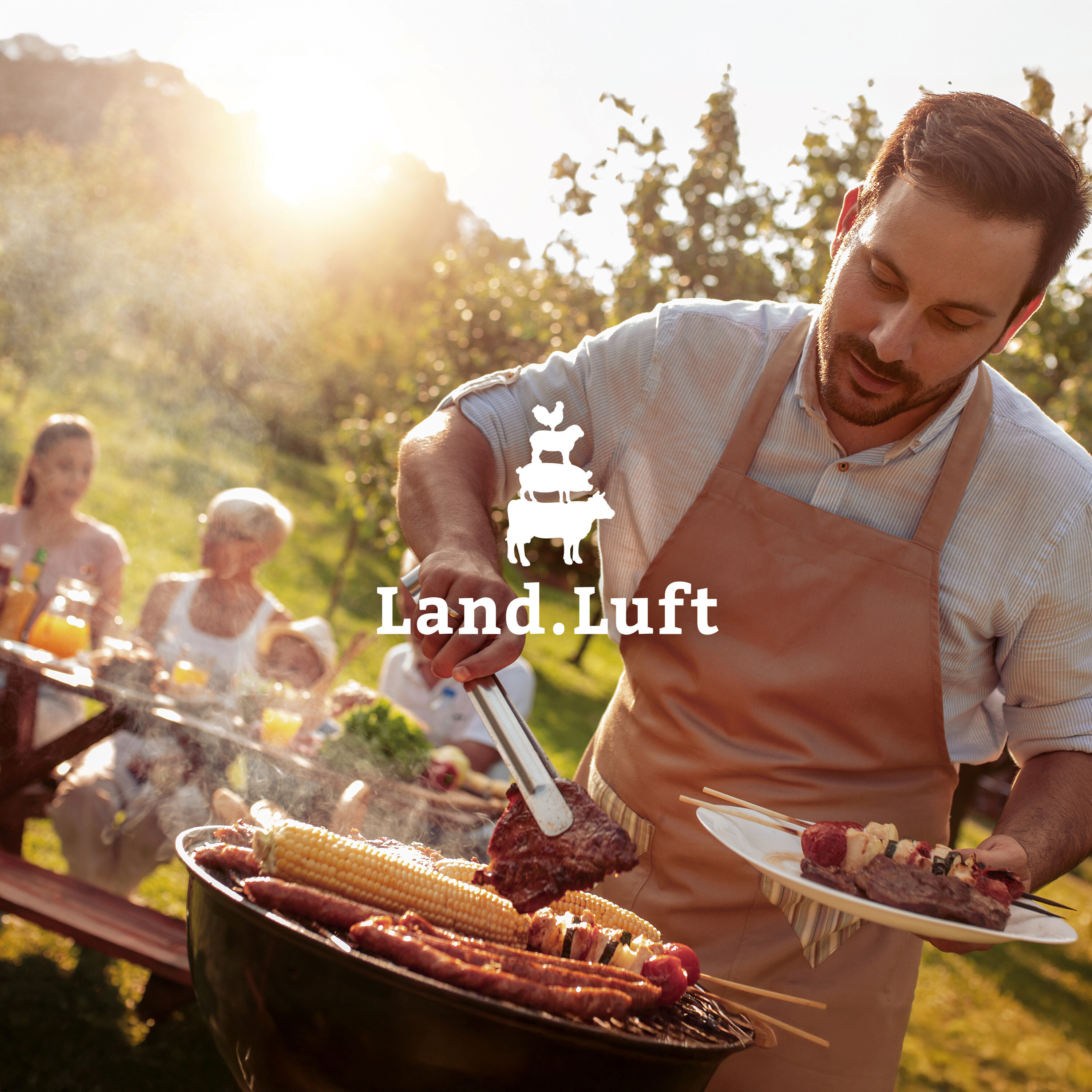 Mann grillt Fleisch und Gemüse im Freien, während Familie am Tisch sitzt. Logo von Land.Luft im Vordergrund.