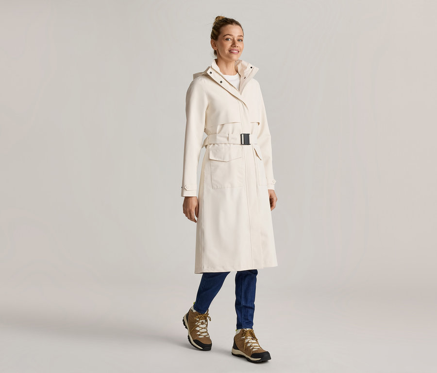 Frau in einem langen, cremefarbenen Mantel mit Gürtel, blauen Jeans und braunen Stiefeln.
