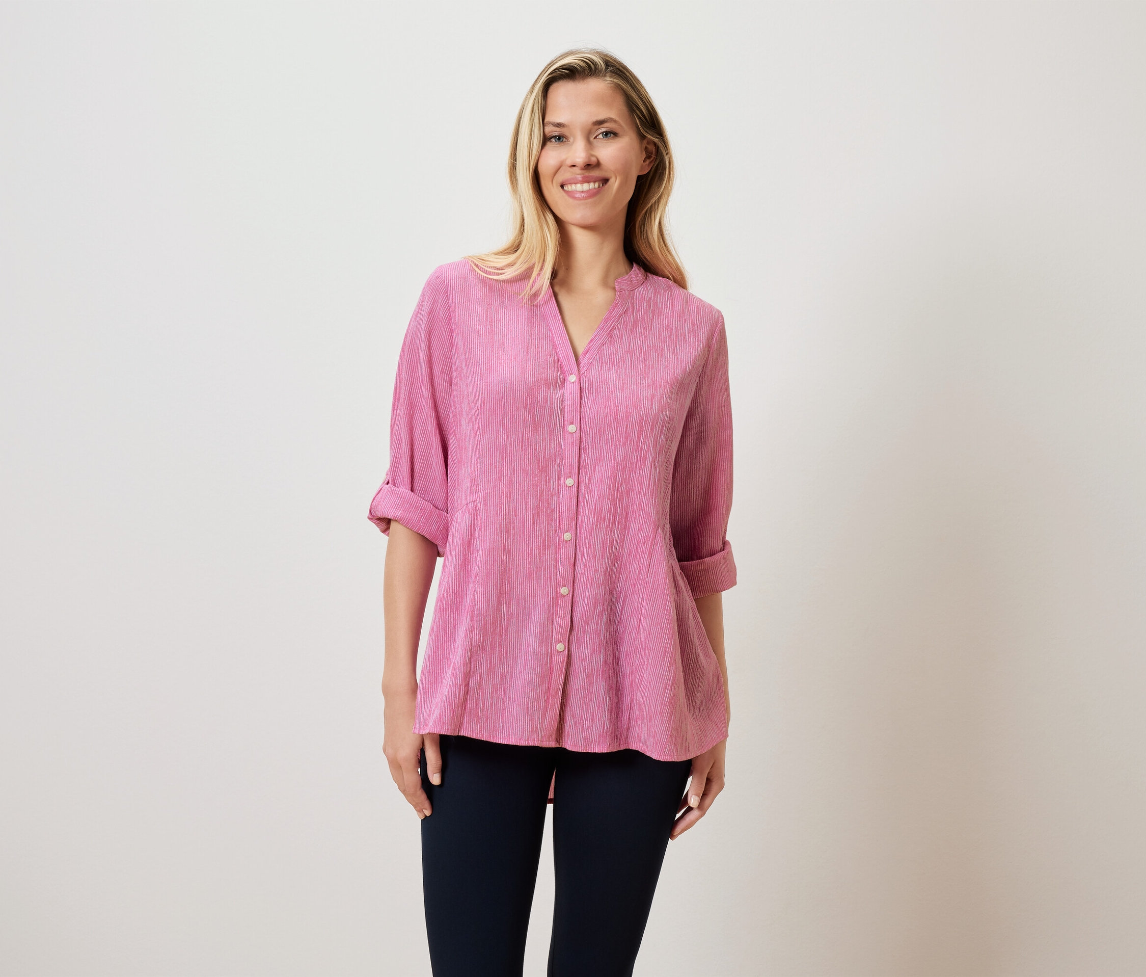 Eine Frau mit blonden Haaren trägt eine pinke Bluse und dunkle Hosen.