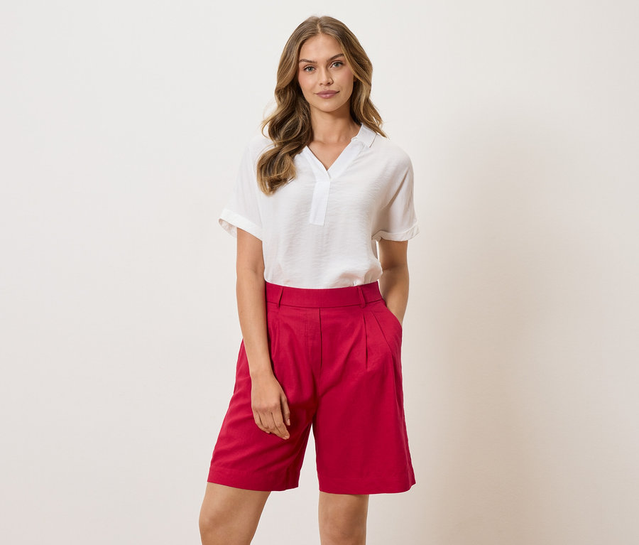 Frau mit weißer Bluse und roten Shorts steht vor einem hellen Hintergrund.