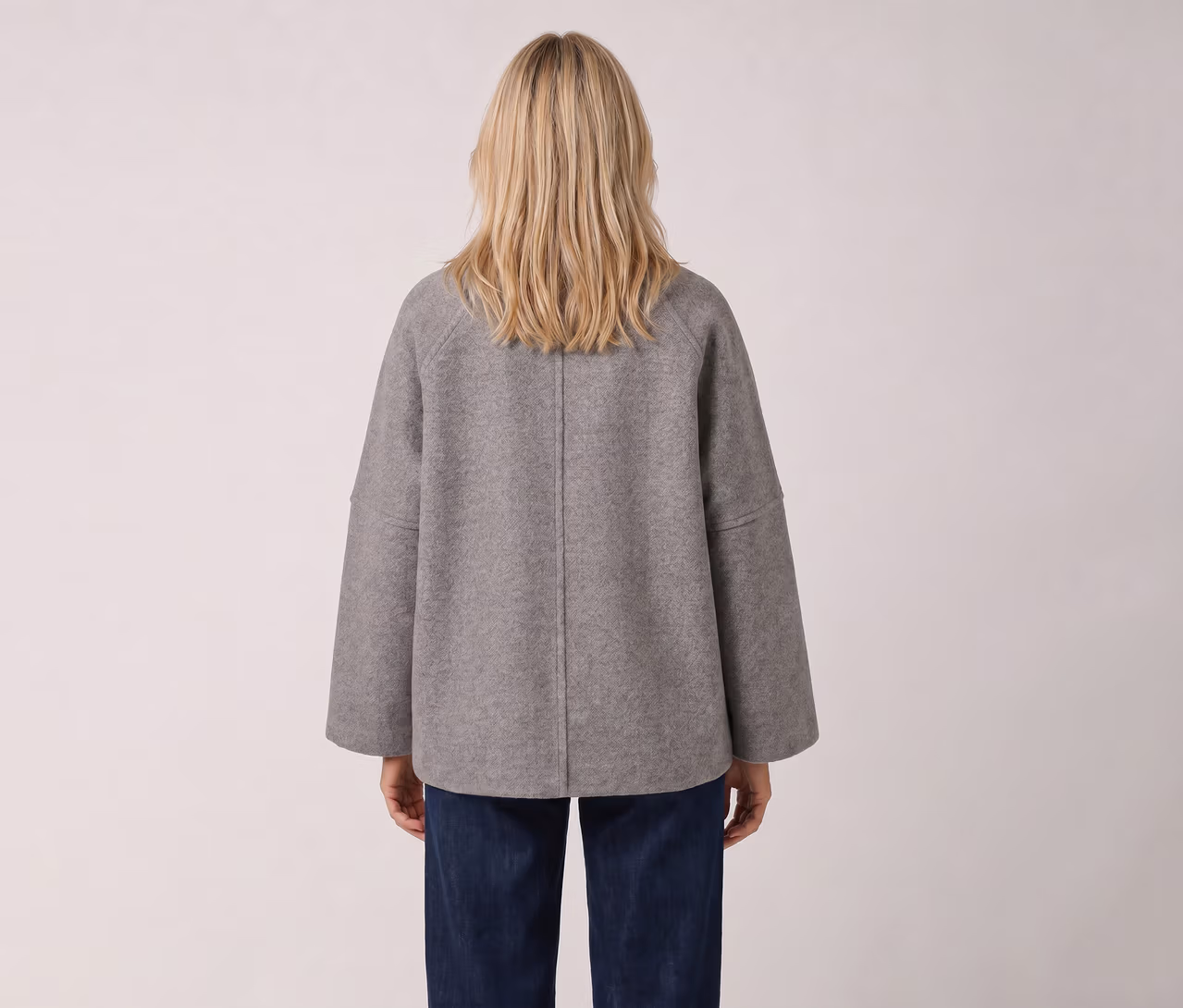 Person von hinten, mit blondem Haar, trägt eine graue, locker sitzende Jacke und blaue Jeans.